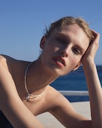 Placeholder for: A person poses outdoors near a body of water, wearing a silver necklace, with their hand resting on their head against a clear blue sky.