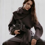 Placeholder for: A person wearing a thick, dark wool sweater with a scarf and holding a matching handbag, sitting in front of a light background.