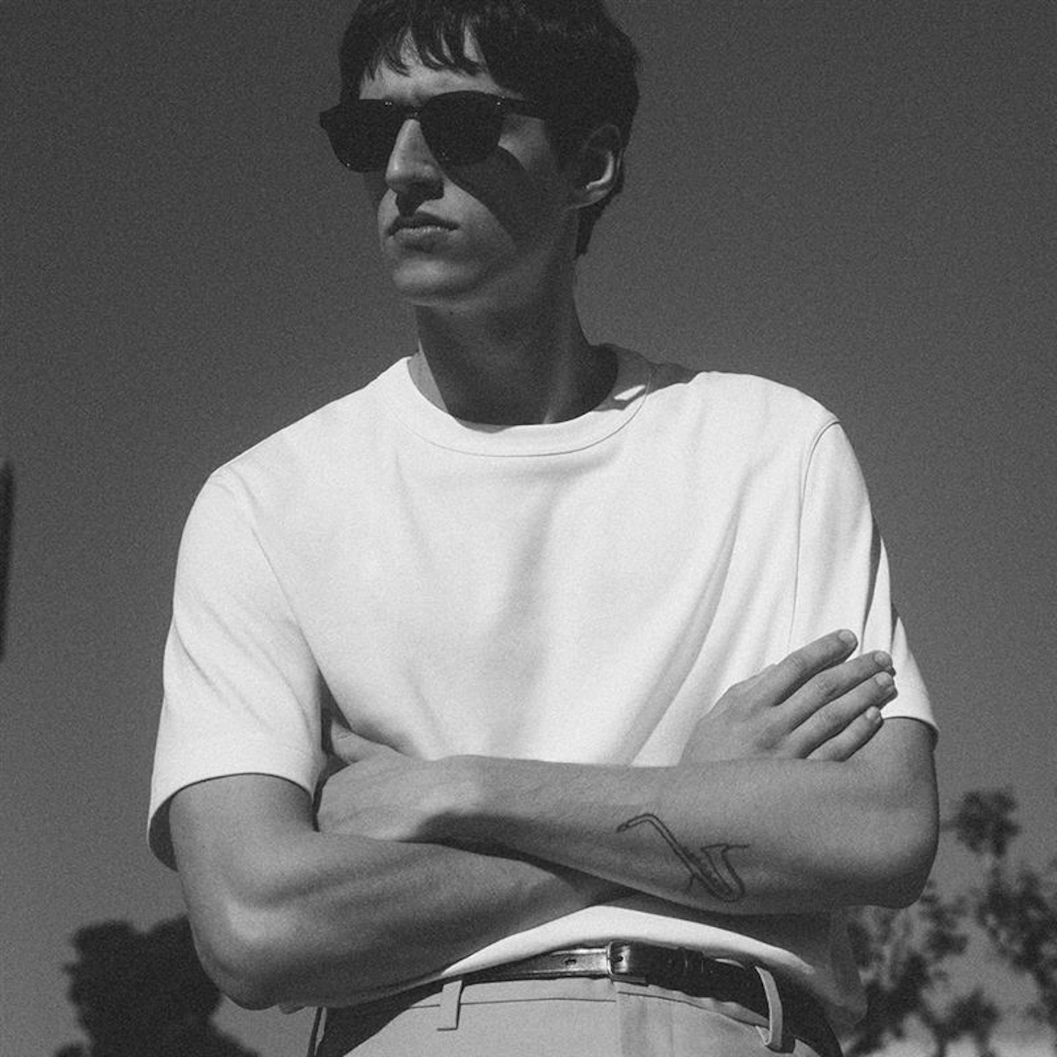 Man in sunglasses and white t-shirt stands with arms crossed. Tattoo visible on forearm. Black and white image with blurred background.
