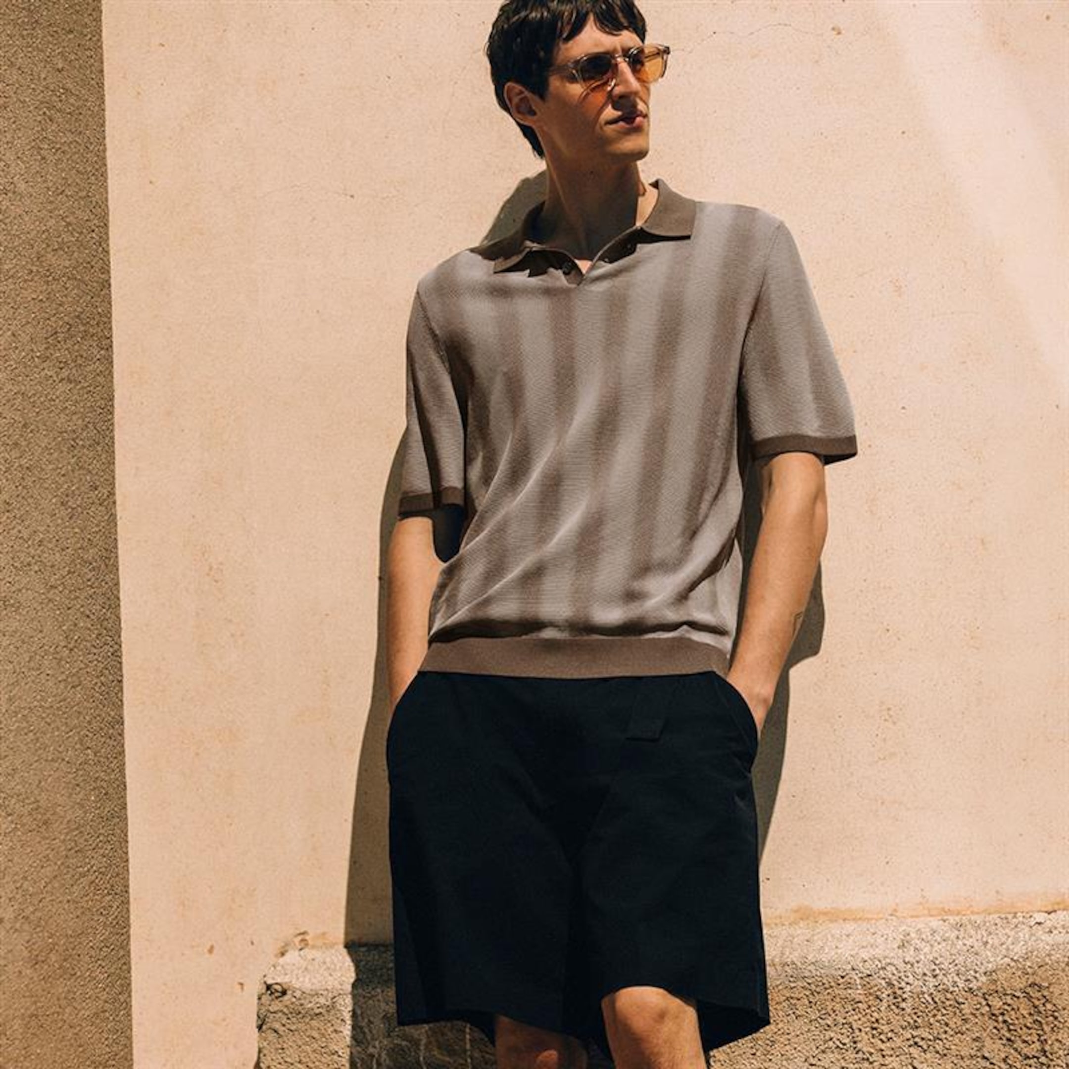 Man in sunglasses wearing a striped polo shirt and black shorts, leaning against a sunlit beige wall.