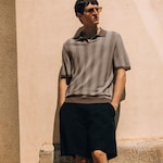 Placeholder for: Man in sunglasses wearing a striped polo shirt and black shorts, leaning against a sunlit beige wall.