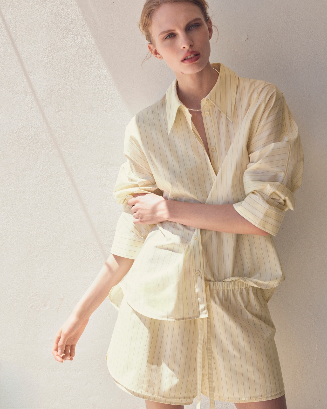 Person in striped yellow shirt dress leans against a sunlit white wall, looking towards the camera with a relaxed expression.