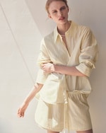 Placeholder for: Person in striped yellow shirt dress leans against a sunlit white wall, looking towards the camera with a relaxed expression.