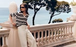 Placeholder for: A woman in sunglasses leans on a balcony wearing a striped top and cream pants, with the ocean and a tree in the background.