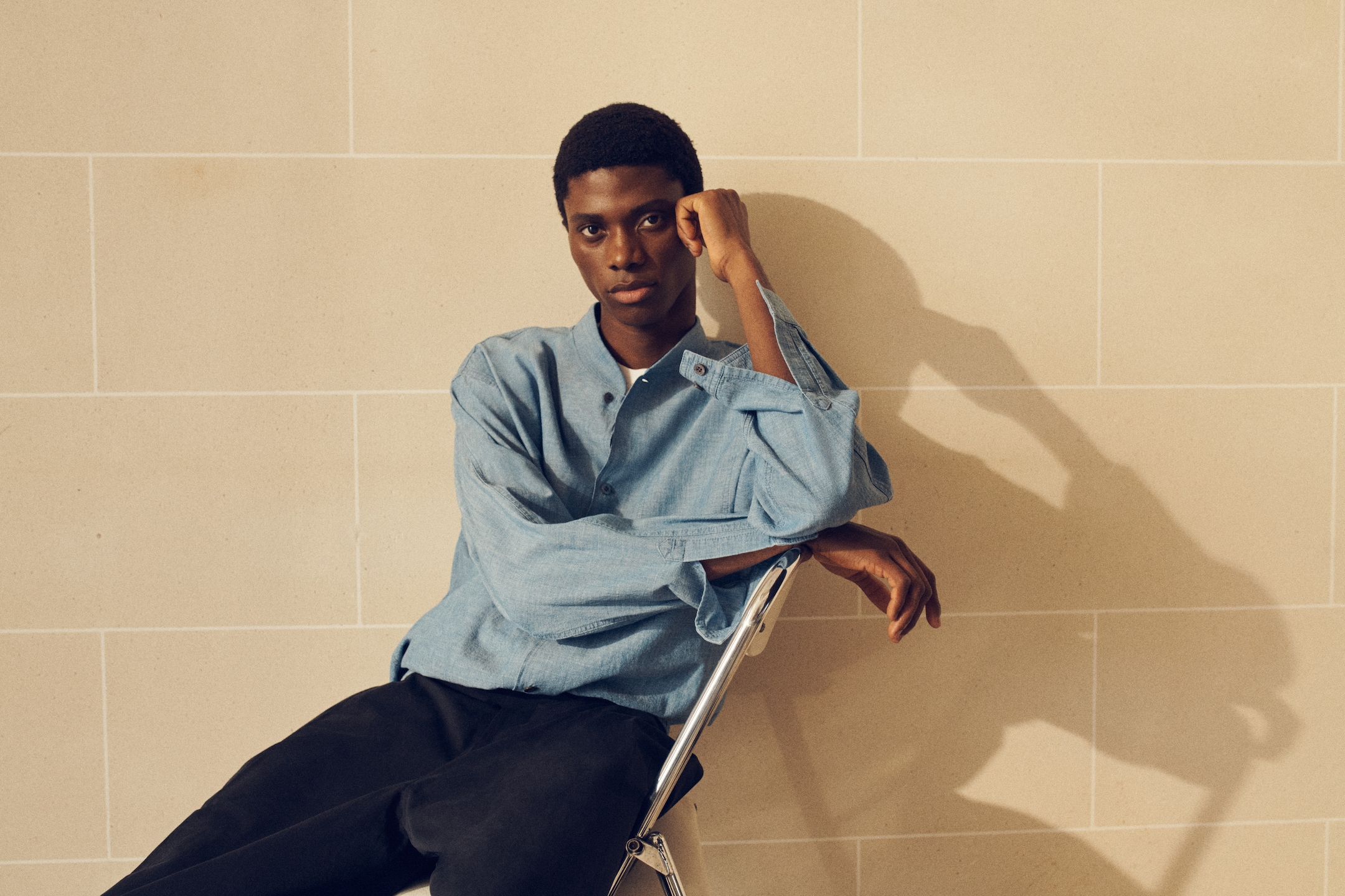 A person in a light blue shirt sits thoughtfully on a chair against a beige tiled wall, casting a shadow.
