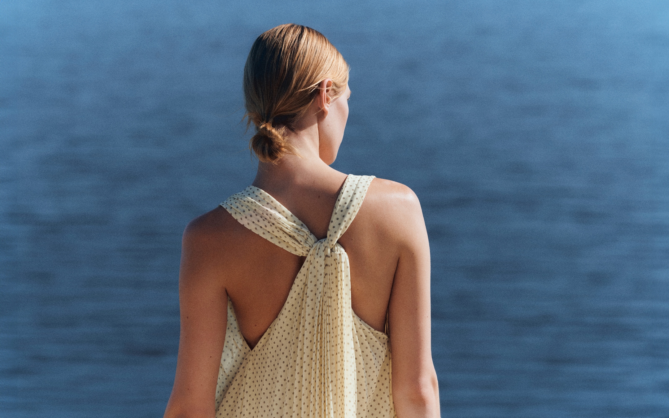 Woman in a sleeveless dress with a back knot stands overlooking calm blue water.
