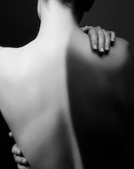 Placeholder for: Black and white photo of a person's bare back with hands resting on the shoulder, casting dramatic shadows.